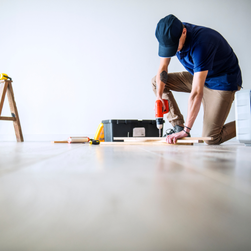 A home remodeling worker drills into a wooden floor, focused on his task in a partially renovated room.