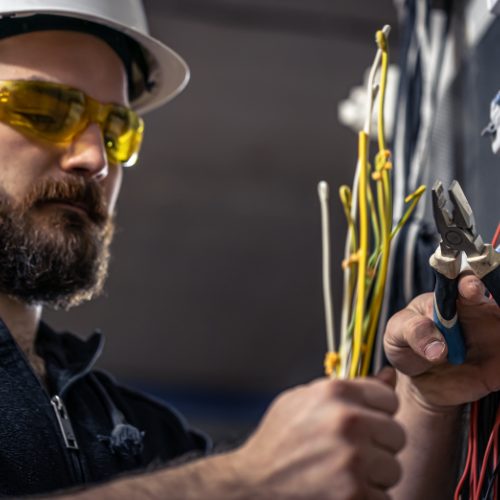 A man with glasses and a beard, dressed as an electrician, is holding a wire, ready to perform electrical tasks.