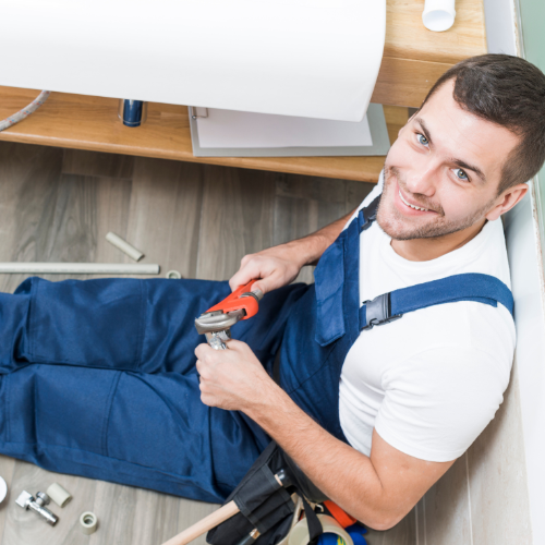  A man in overalls sits on the floor with various plumbing tools, preparing for a task.